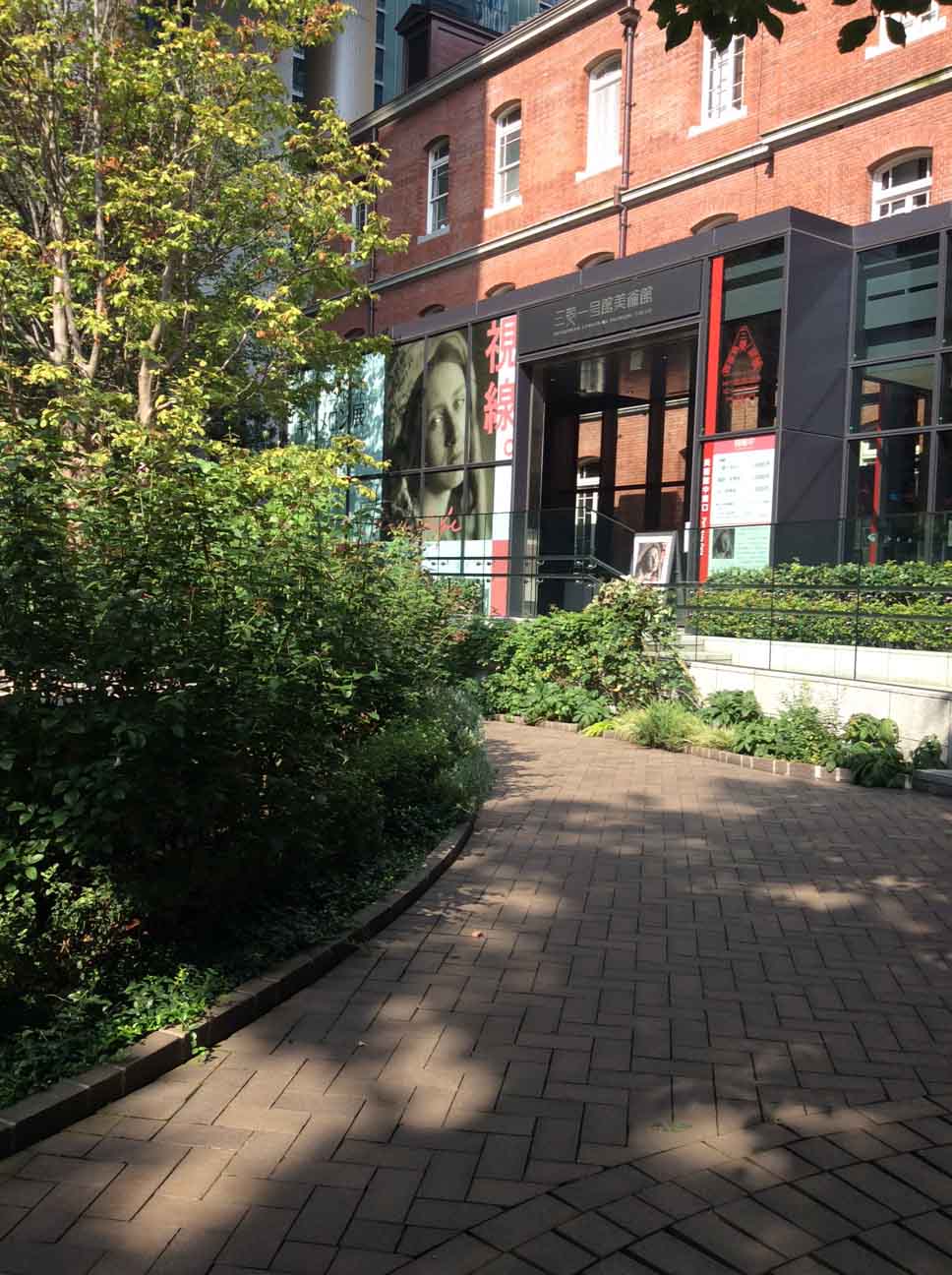 a red-brick path curves left in front of the museum entrance, greenery on the left of the picture partially obscures the poster of Julia Margaret Cameron on the red brick wall of the Museum.